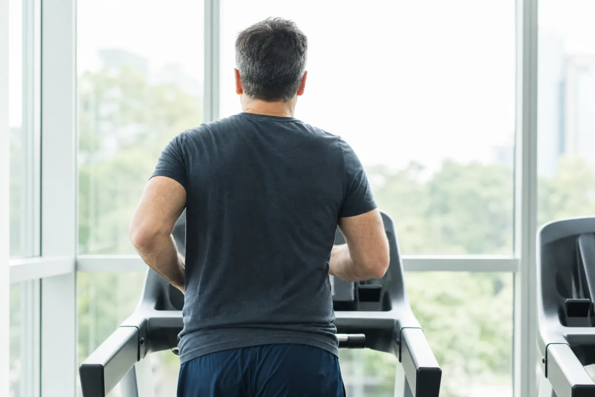 A man running on a treadmill beside floor-to-ceiling windows in a bright gym, photographed from behind