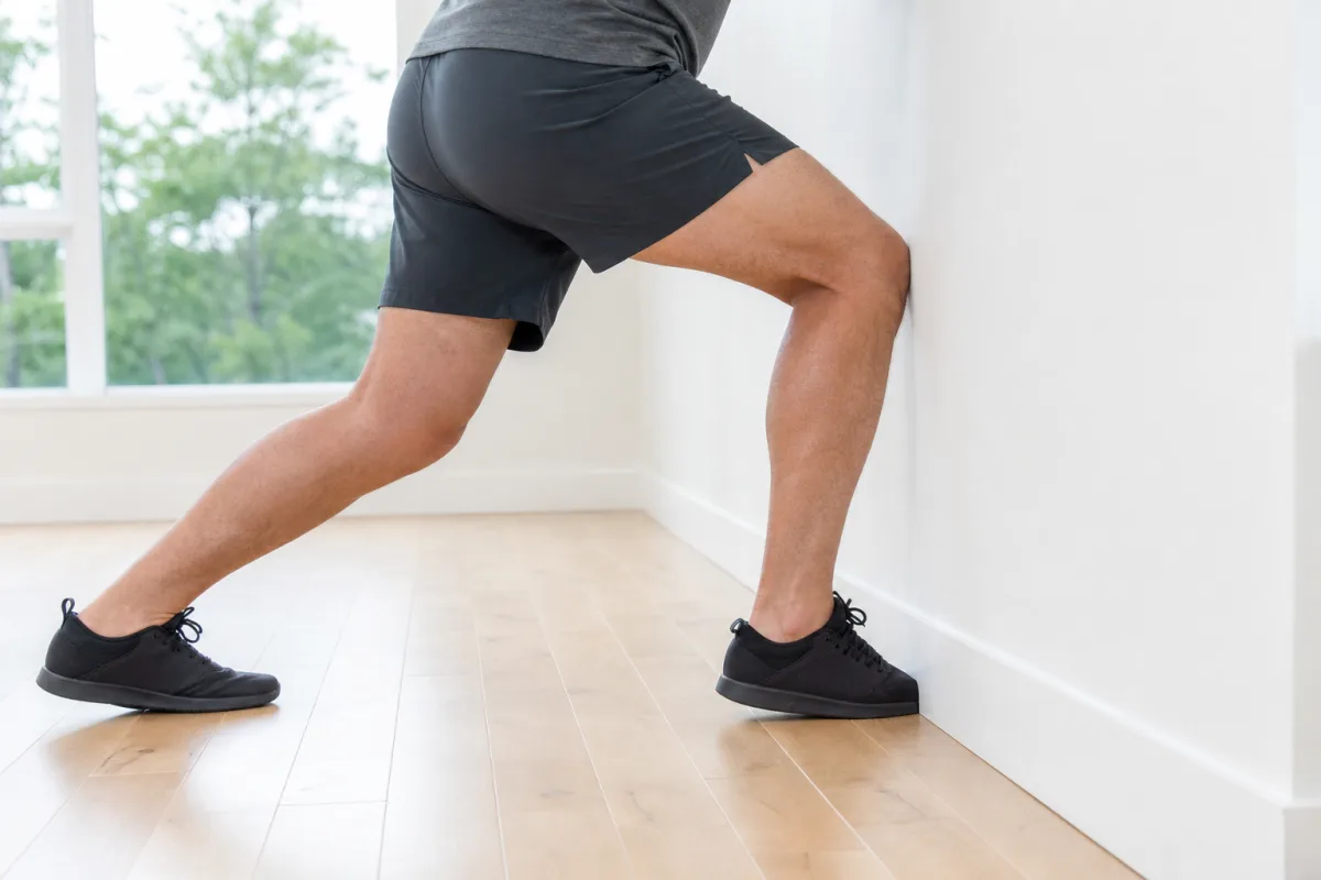 A person's legs in a wall-supported ankle position with the knee driving forward toward the wall, on a bright wooden floor