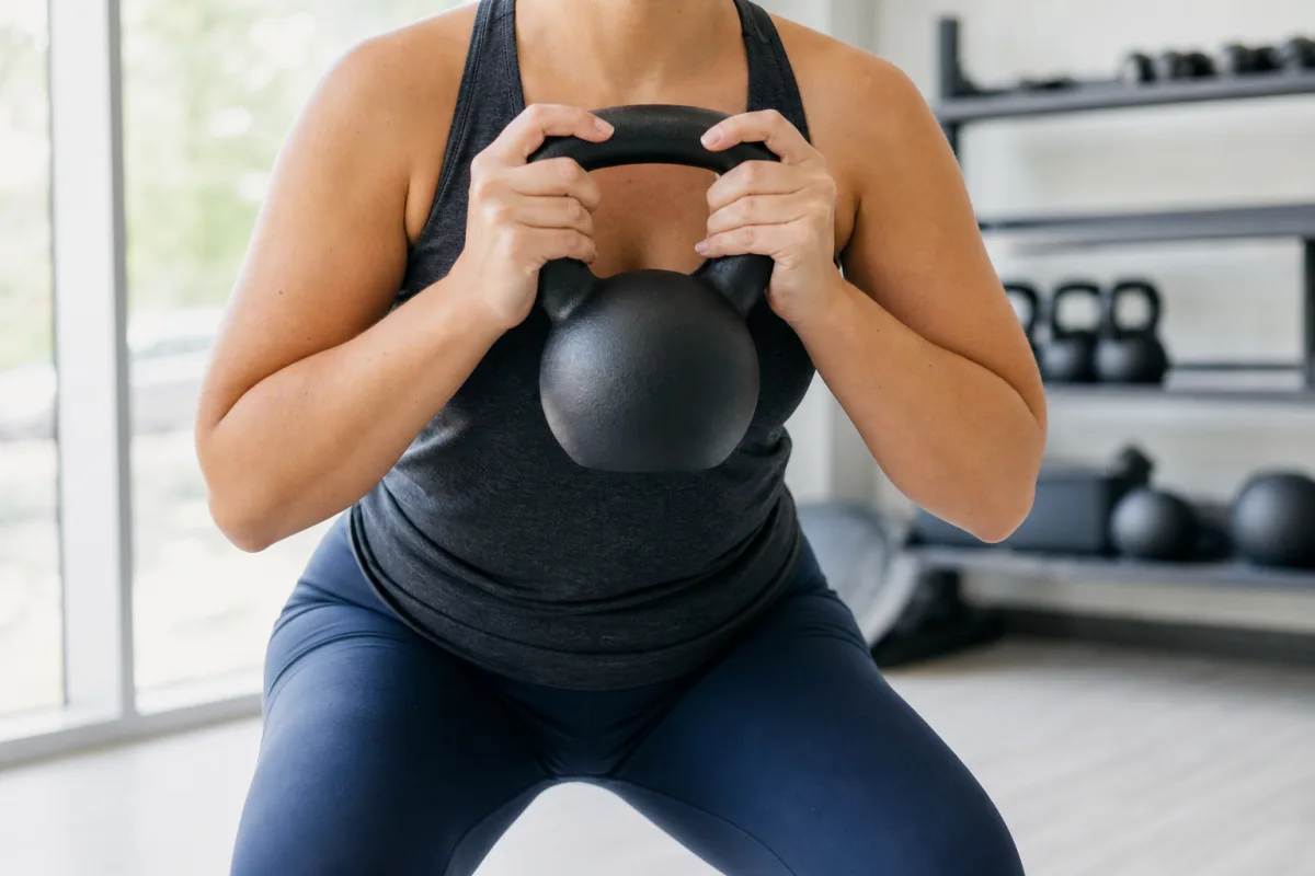 A woman holding a kettlebell at chest height in a goblet squat position in a bright gym